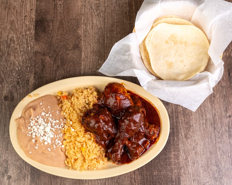 Chicken mole with rice, refried beans, and a side of tortillas on a wooden table.