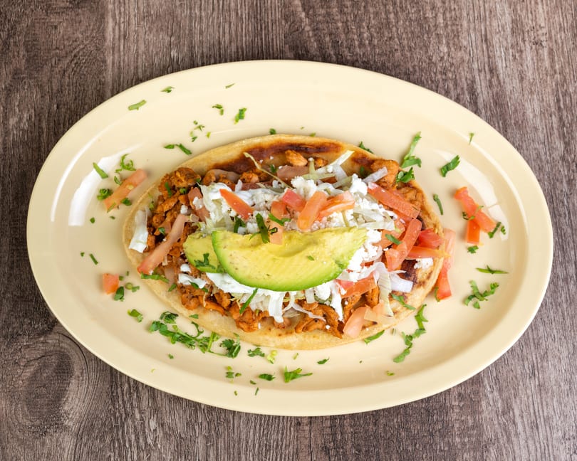 A sopes with chicken, lettuce, tomatoes, avocado, and cheese on a yellow plate.