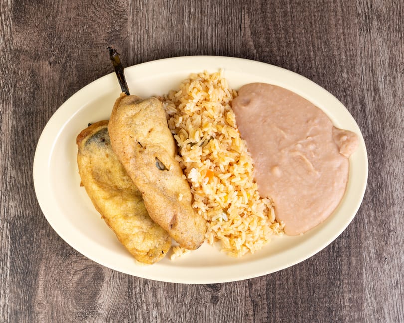 Two chile rellenos, rice, and refried beans on an oval plate on a wooden table.