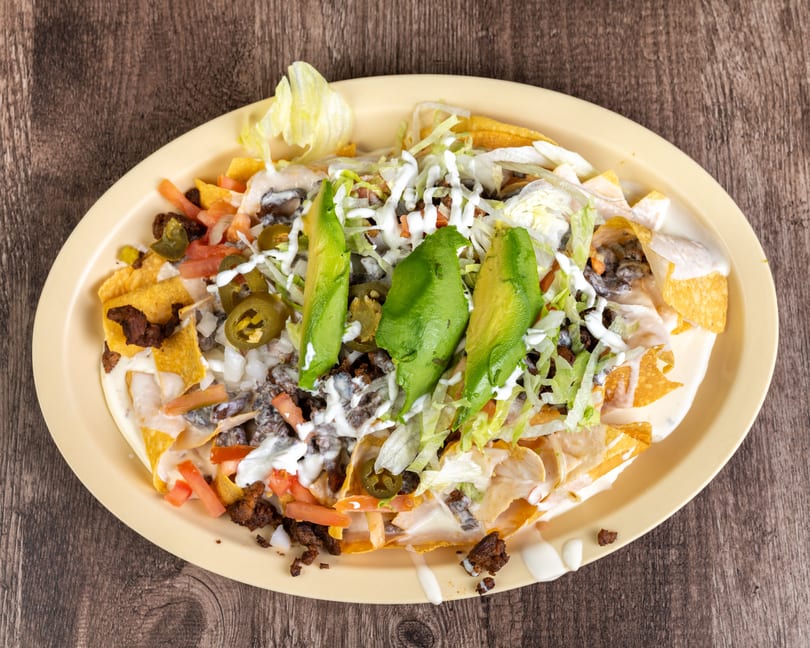 A plate of nachos with ground beef, cheese sauce, lettuce, tomatoes, jalapeños, and avocado slices.