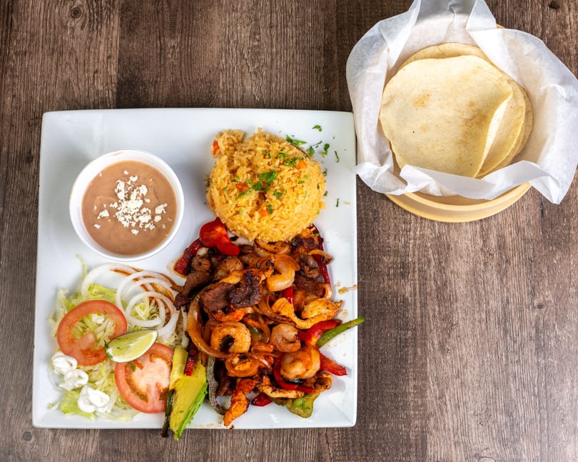 A fajita platter with shrimp, steak, chicken, rice, beans, salad, and tortillas on a wooden table.