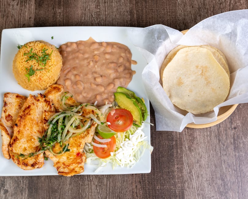 Grilled chicken with rice, refried beans, salad, avocado, and tortillas on a wooden table.