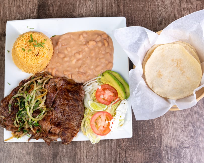 Grilled steak with onions, rice, refried beans, salad, avocado, and tortillas on a wooden table.