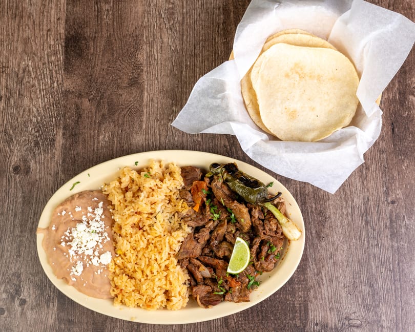 Carne asada plate with rice, refried beans, grilled pepper, and a side of tortillas.