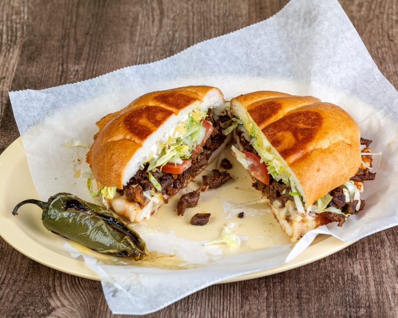 A torta sandwich, cut in half, filled with meat, lettuce, tomato, and cheese, served with a grilled jalapeño.